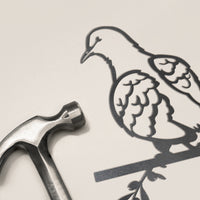A metal hammer sits beside the Turtle Dove, a slim and elegant Corten steel bird ornament showing a dove on a branch, both set on a light surface.