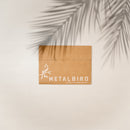 A brown cardboard box with the METALBIRD logo, bird illustration, and “Proudly Made Here in America.” The Hummingbird & Chicks metalbird is a thoughtful Mother's Day gift. A palm leaf shadow above adds a natural touch.