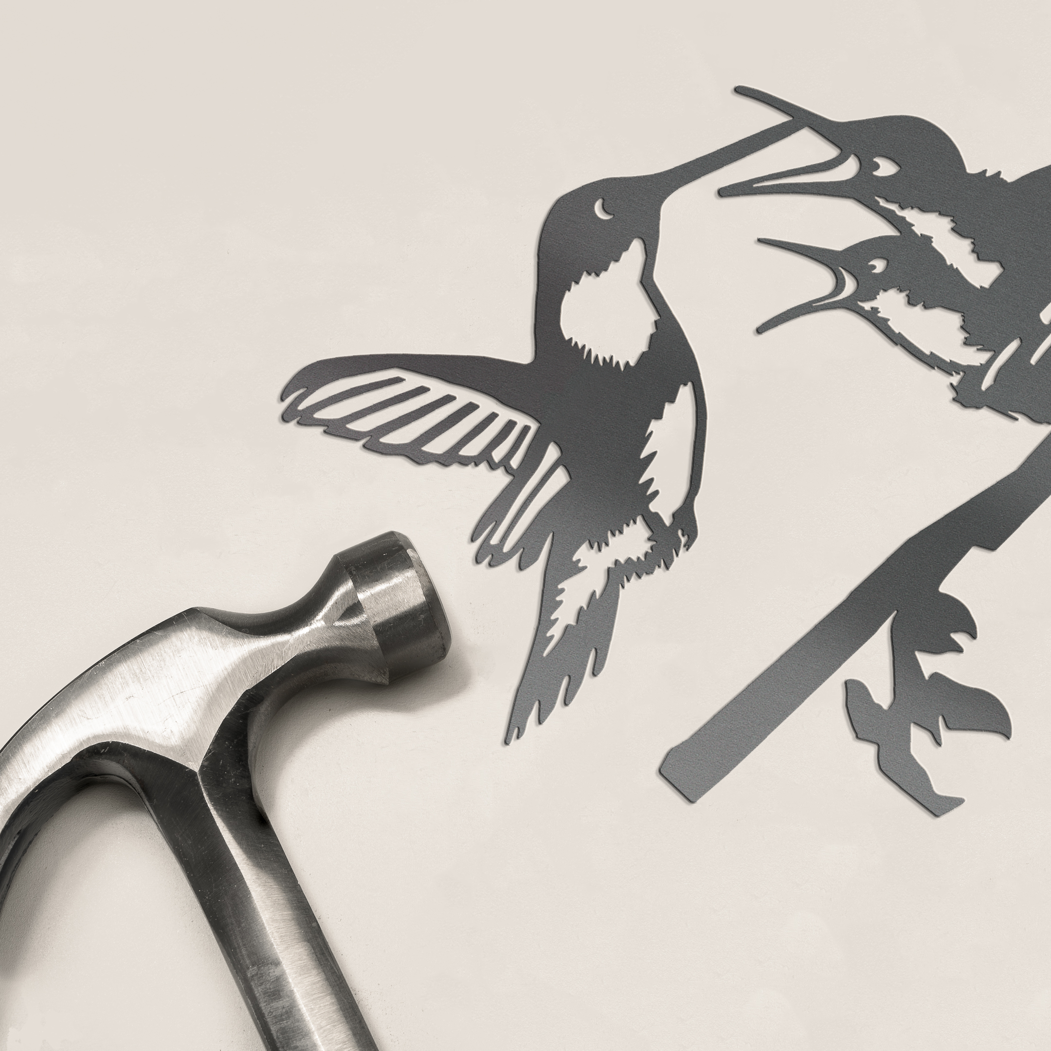A silver hammer lies beside the Hummingbird & Chicks metalbird silhouette—two corten steel birds on a branch, facing each other—a charming decor piece and perfect Mother’s Day gift.