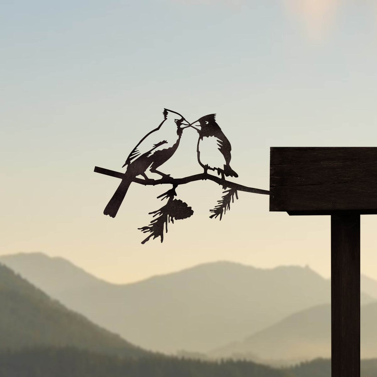 Two kissing cardinal silhouettes mounted on a timber rail against a mountain view.