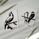 The Metalbird Greeting Card Set features two white cards with black bird silhouettes—one with a single bird on a branch, the other showing two birds on a wire. Surrounded by green plant stems, these cards make a thoughtful display or gift.