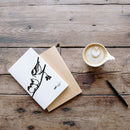 The Metalbird Greeting Card Set, featuring a bird silhouette design, comes with a brown envelope and is displayed alongside a black pen and a cup of latte with heart-shaped foam on a rustic wooden table.