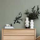 A wooden dresser with stacked books, ceramic cups, and vases stands by a green wall. Above it, the Cardinal Wallbird matte black wall decor shows a cardinal perched on a branch next to two white vases with greenery.