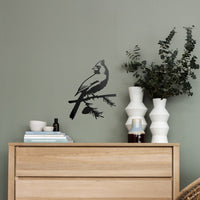 A wooden dresser with stacked books, ceramic cups, and vases stands by a green wall. Above it, the Cardinal Wallbird matte black wall decor shows a cardinal perched on a branch next to two white vases with greenery.