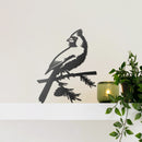 The Cardinal Wallbird, a matte black bird silhouette wall decor, sits on a white shelf near a green candle and leafy potted plant, set against a plain light-colored wall.