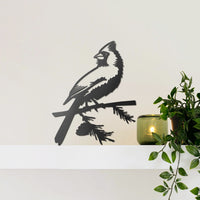 The Cardinal Wallbird, a matte black bird silhouette wall decor, sits on a white shelf near a green candle and leafy potted plant, set against a plain light-colored wall.