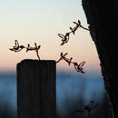The Baby Bird Bundle - Hummingbird Trio features three mini hummingbird silhouettes and flower ornaments crafted from Corten steel, displayed on a wooden post and a tree, with a soft sunset sky in the background.